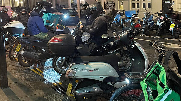 Motorbikes parked up in a row on a road at night, text says "Tell RBKC to sort out the delivery bikes!"
